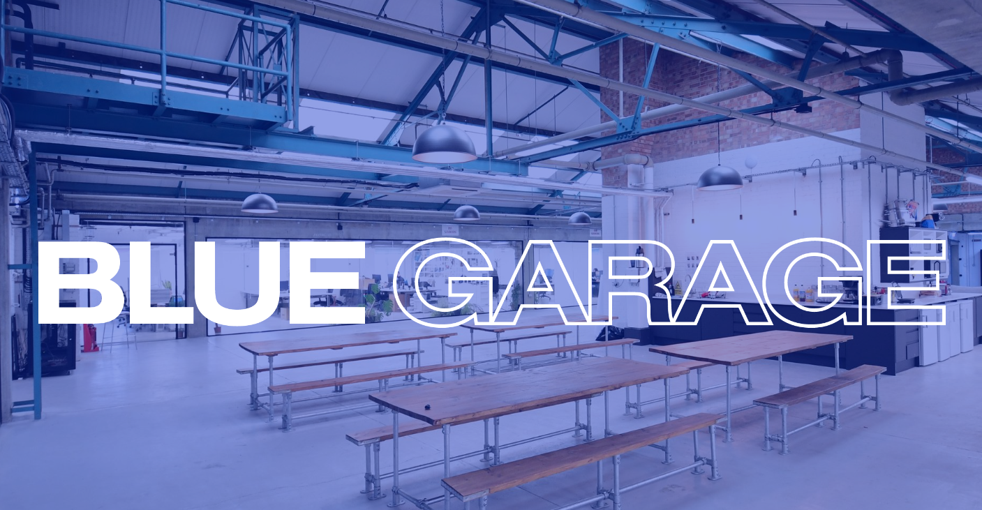 The interior of a large, industrial-style space with a blue color overlay. Long wooden tables and benches are arranged in rows under a high ceiling with exposed blue steel trusses. In the foreground, the words